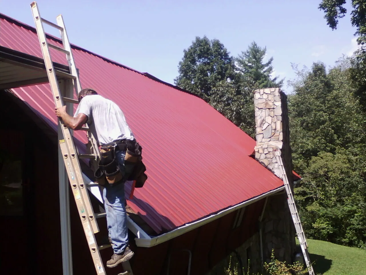 Roofer on a ladder beside a red metal roof and stone chimney — Soffit & Fascia Repair by Roofing Brothers in Leighton